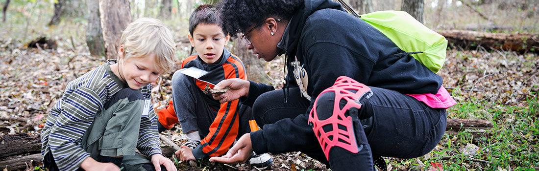 A teacher with two young children are inspecting a leaf in the woods
