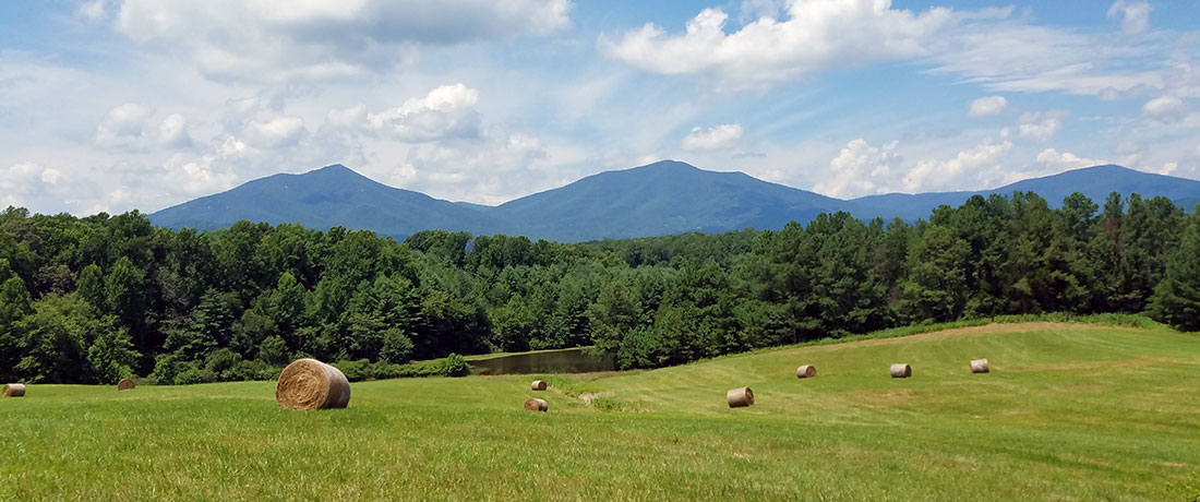 An open field with hay bales, and a pond, forest, and mountains in the background